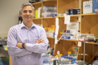 A man stands with arms folded, in front of shelves containing laboratory equipment.