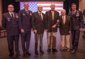 Six men stand elbow-to-elbow on a stage with a large American flag projected behind them