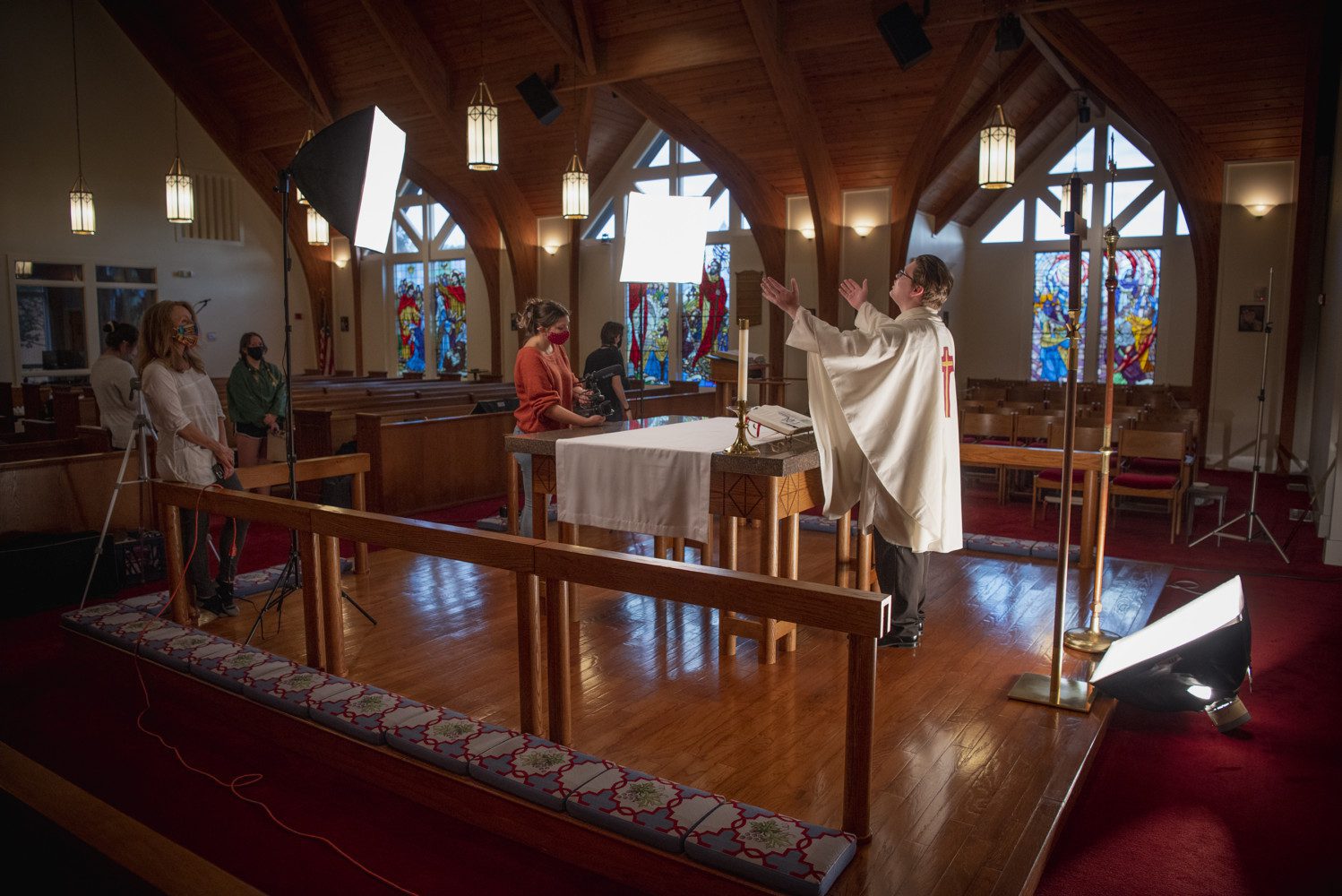 Several people wearing masks stand in a church under a lighting set-up, a man playing a preacher rehearsing in the foreground