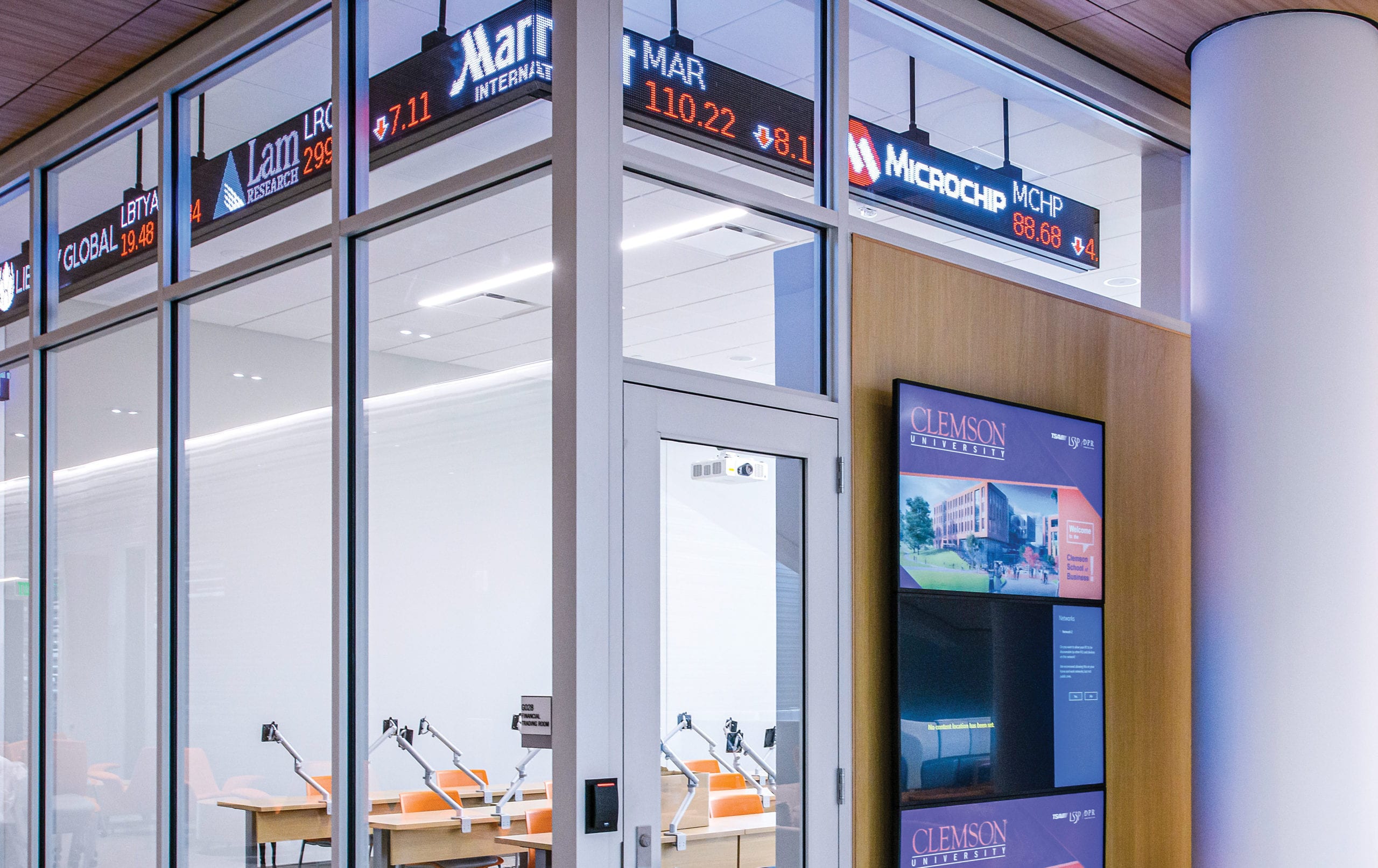 A glass and steel classroom with a wood panel inset on the outside that holds a digital screen for messages. An electronic stock ticker display wraps around the front and glass side of the classroom, displaying logos and stock information. Visible from the glass wall, the inside of the classroom has long desks and chairs with patterned carpet.