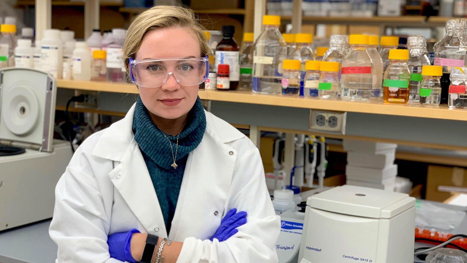 Woman wearing lab coat, safety goggles and purple gloves standing in a lab with her arms crossed