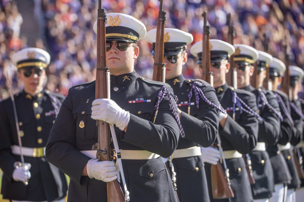 A formation of people in formal military uniforms stand at attention holding rifles in front of them.