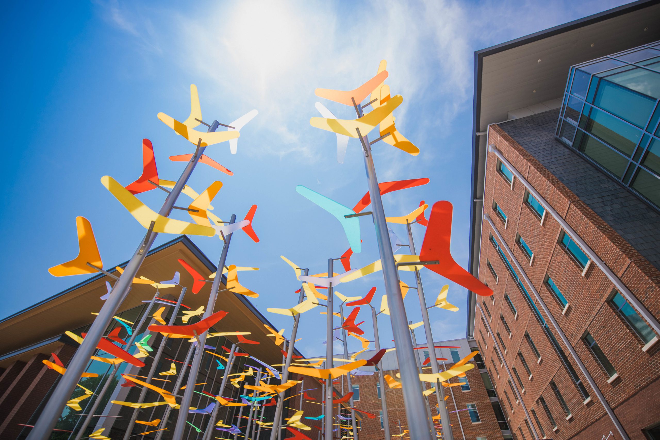 The sun shine down through dozens of colorful boomerang-shaped shapes attached to poles in front of the CORE Campus building