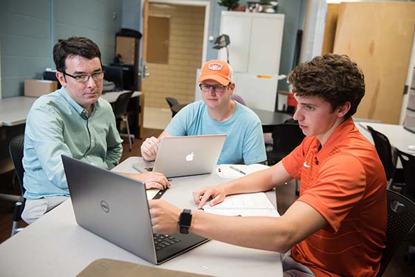 three men sitting around a table and looking at a laptop screen.