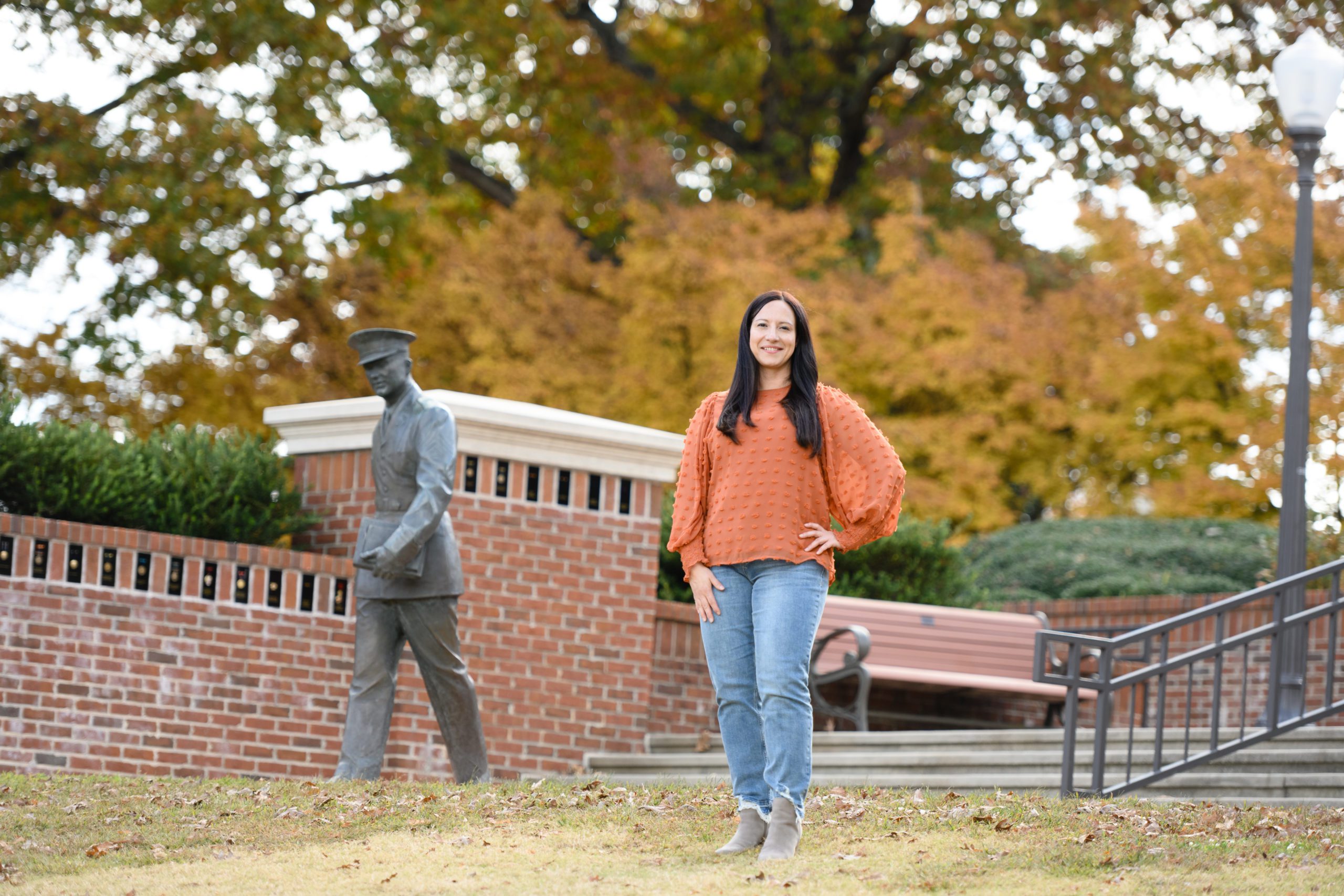 Biological sciences student Rachelle Hales wears an orange sweater and jeans and is standing with her hands on her hips in a plaza with a military statue.