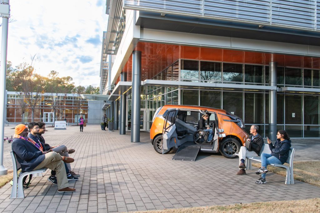 An orange capsule-shaped car has its doors opened, revealing its wheelchair-accessible capabilities. Five individuals sit on two benches near the car.
