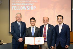 Four men in suits pose together in front of a screen reading "Electron Microscopy Fellowship." The youngest man holds a diploma.