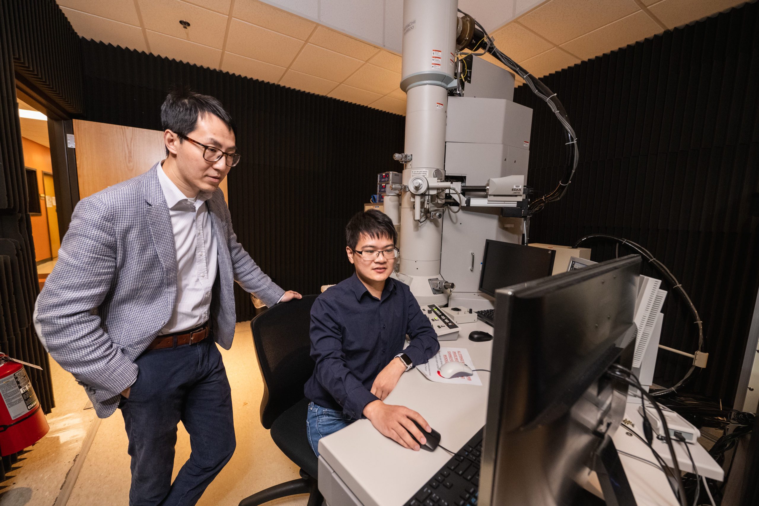 Two men working at a computer next to a large white electron microscope.