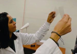 A woman in a white lab coat holds up two squares of grey, biodegradable plastic.