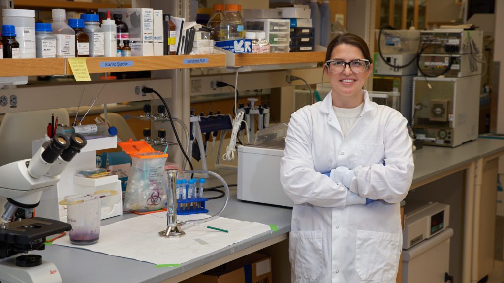 A woman wearing a white lab coat stands in front of a table in a lab at the Clemson University Life Sciences Facility