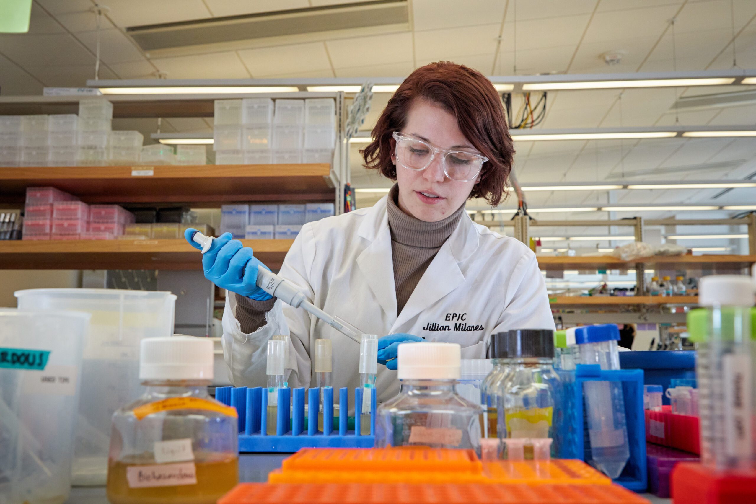 A young woman with red hair wearing a white lab coat and blue plastic gloves holds a pipette that she is using to fill blue tubes.