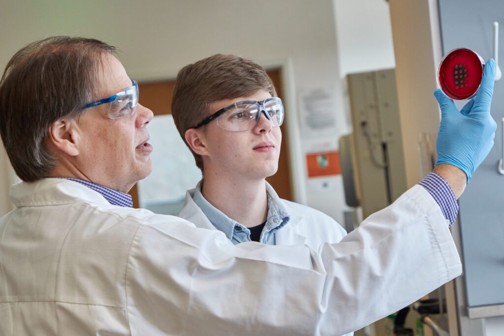 In the foreground, and older man wearing a white lab coat, safety goggles and blue gloves holds up a red petri dish as a younger man also wearing lab gear looks on.