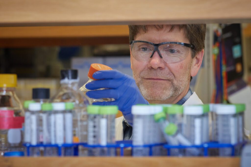 A scientist wearing blue gloves and safety goggles is shown through a pass-through taking blue, green and clear bottles from a shelf.