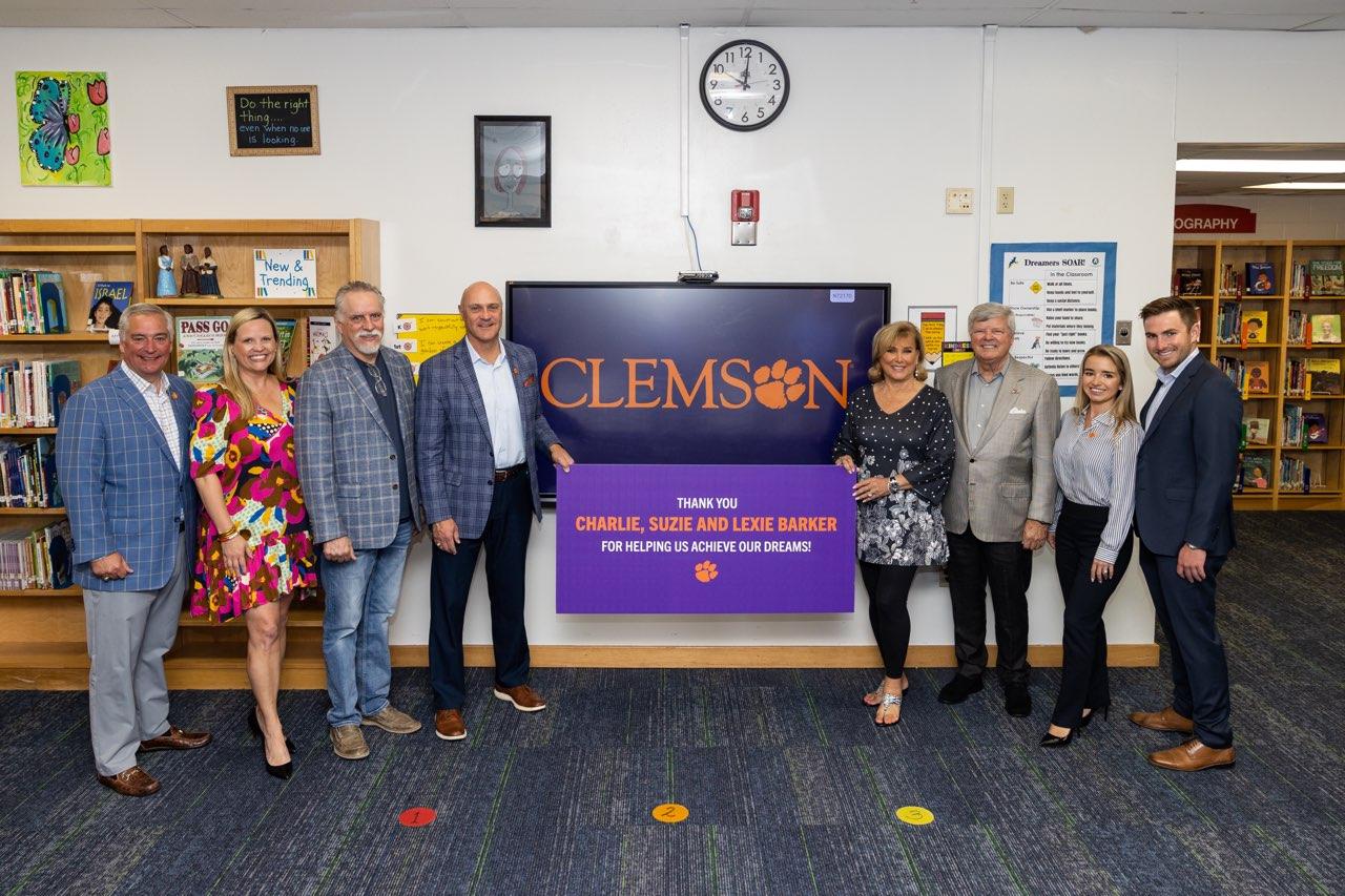 A group of eight people standing in alongside a large wall monitor/tv - four people on each side - in a school library setting. Clemson president and Susan Barker each have an end of a large poster that names the Barker Presidential Scholarship Endowment.