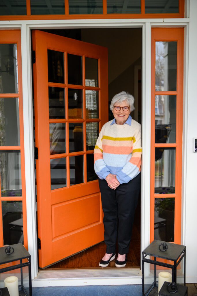 A woman with white hair, wearing glasses and a pastel sweater stands just outside of a slightly ajar orange front door.