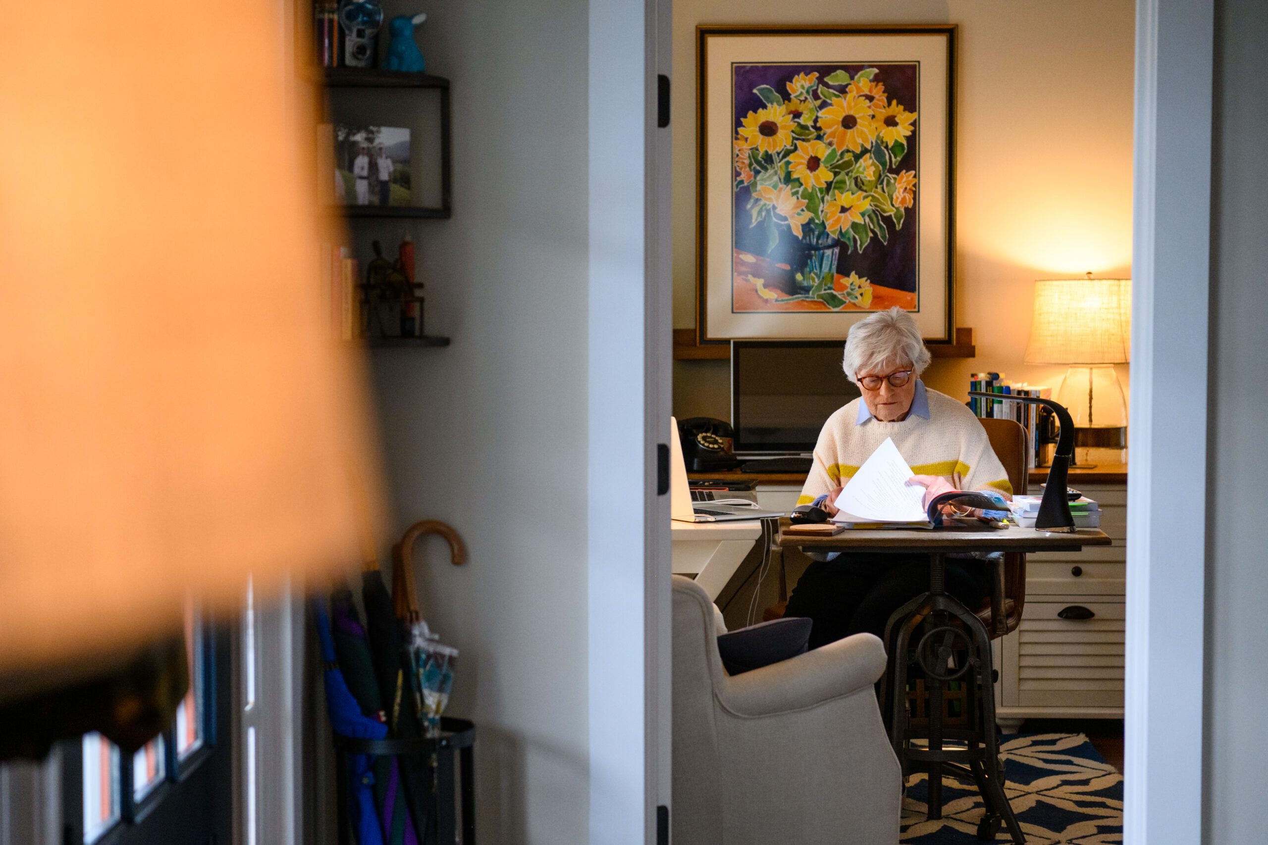 A woman is seen working at a home office desk through an open doorway, flipping through papers.
