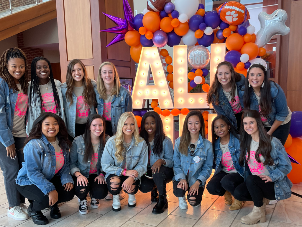 A group of about a dozen college-aged women in denim jackets smile around a large sign that says "All In." Orange and purple balloons adorn the sign. Delaney Toomer is in the front row.