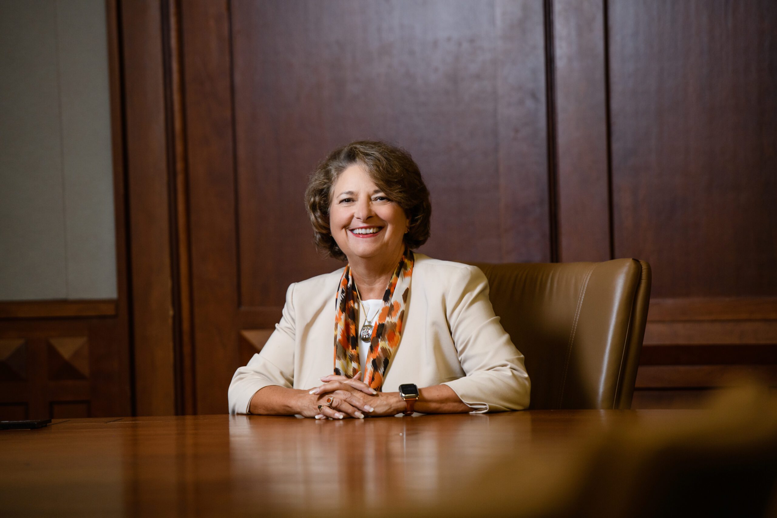 A woman wearing a tiger print scarf and white blazer is seated at a board room table smiling, with her arms folded atop the table.
