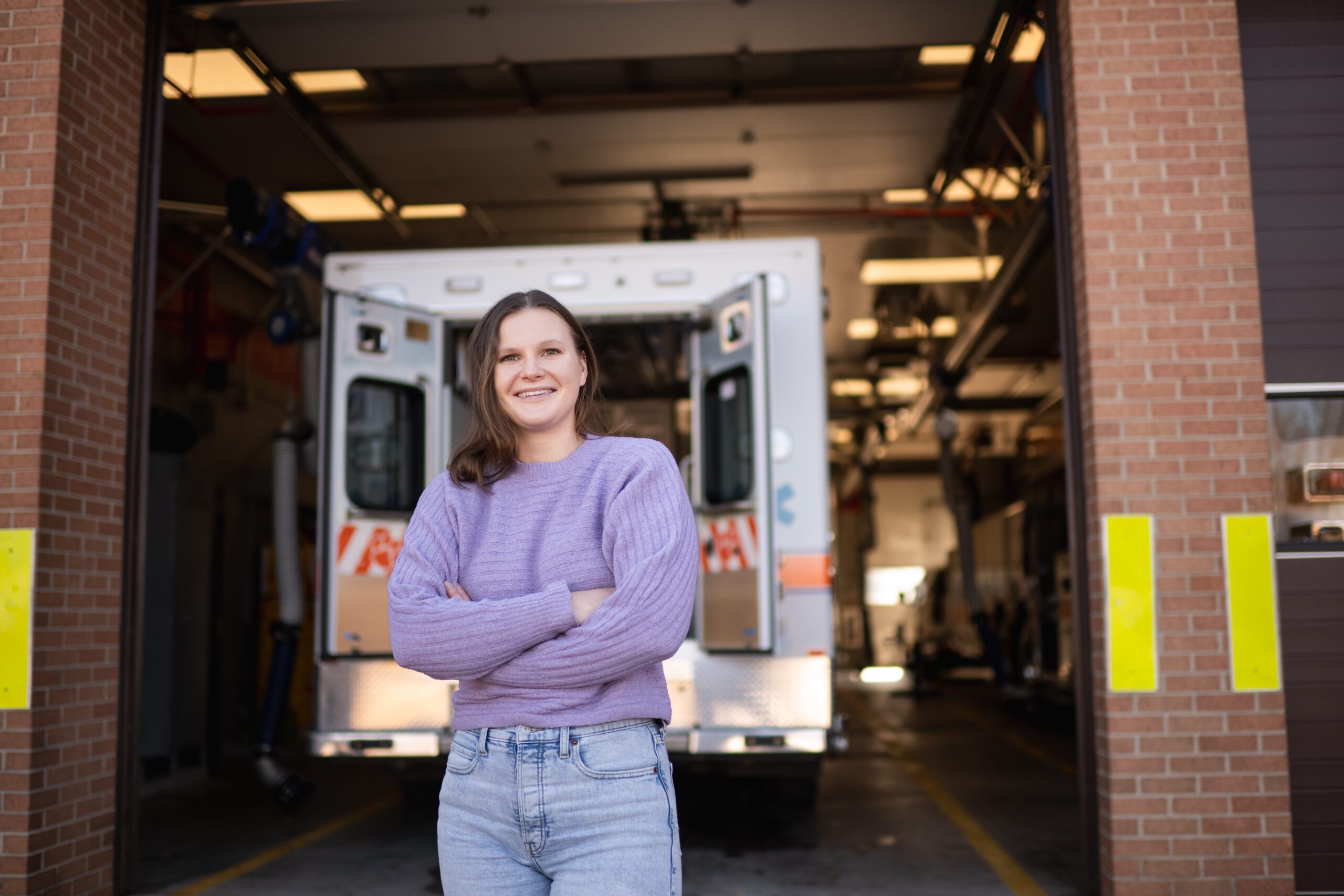 A young woman wearing a purple long-sleeve shirt stands in front of an ambulance bay. Her arms are crossed in front of her.