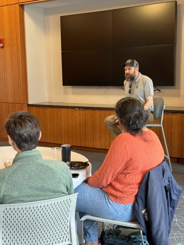 A man seated in a chair speaking to a room full of students.