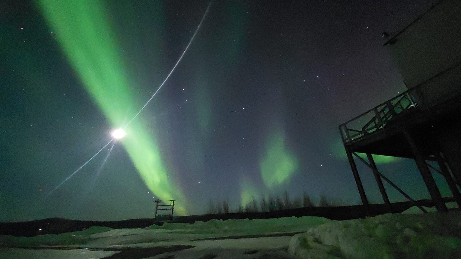 An aurora with a tree line and a building in the foreground.