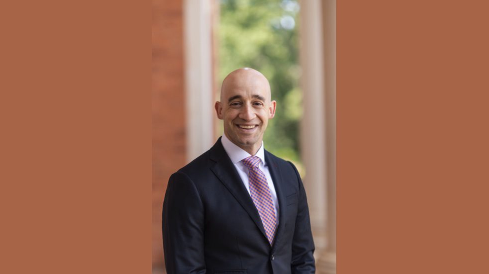 A man dressed in a business suit stands smiling in front of a building.