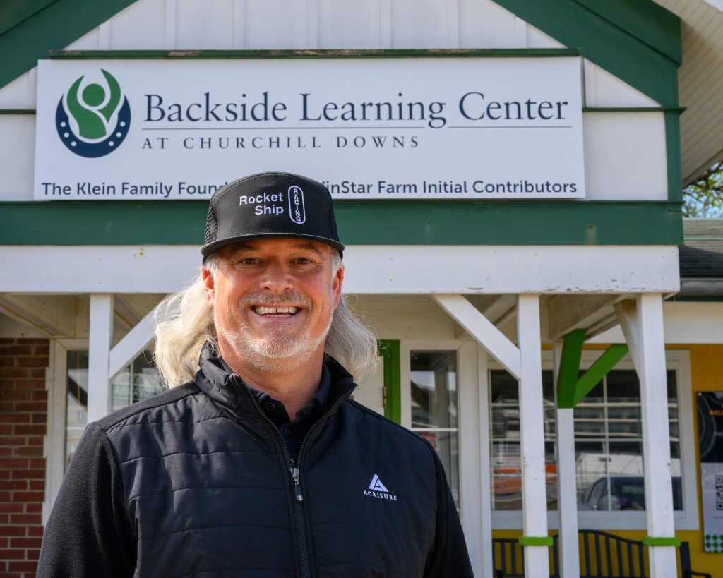 A man with shoulder-length silver hair wearing a black ball cap and a black athletic jacket smiles in front of a sign that reads "Backside Learning Center."