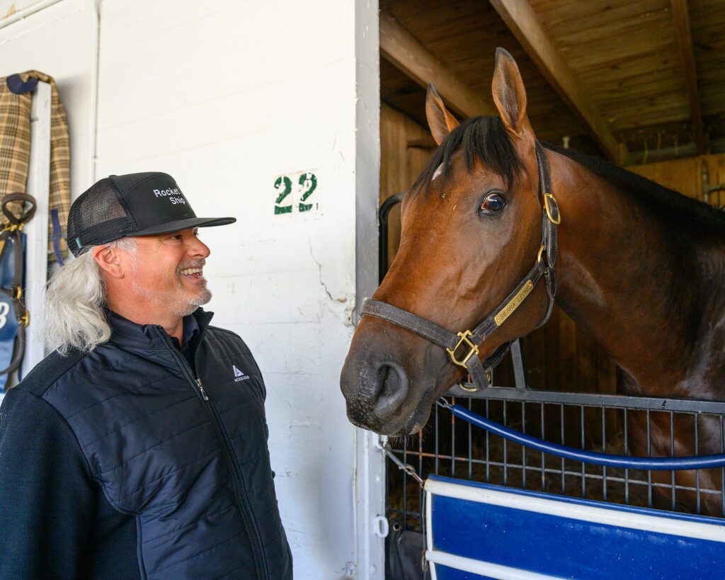 A man with shoulder-length silver hair wearing a black ball cap and a black athletic jacket smiles at large brown a horse looking out from inside a stall.