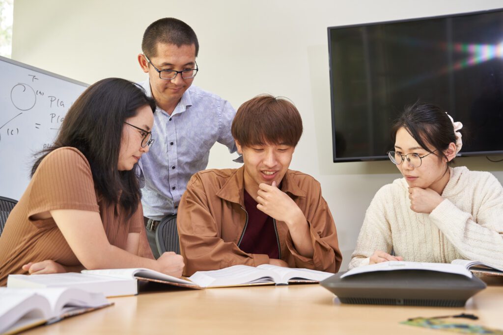A man looks over the shoulder of a man and two women sitting at a table looking at a book
