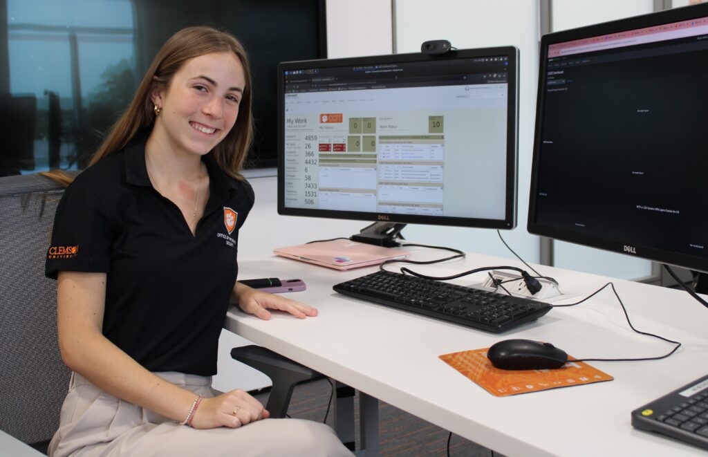 A woman in a black polo sits beside a computer, smiling at the camera