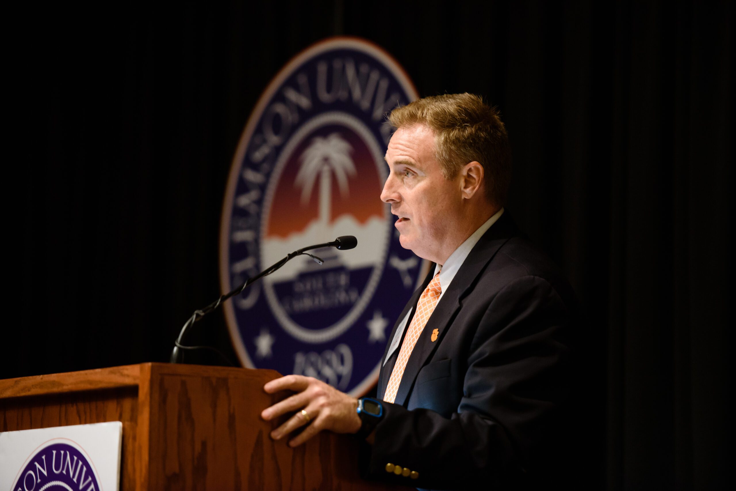 Caucasian man is a suit speaks while standing at a podium. A Clemson seal is in the background.