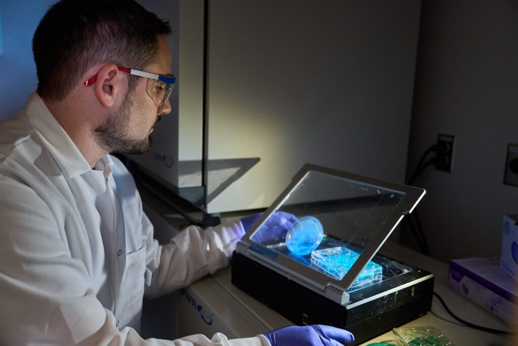 Man wearing a lab coat looks at some Petri dishes lit up by blue lights.