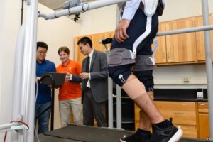 Three men look at a clipboard in the background while someone where a knee brace type device walks on a treadmill in the foreground.