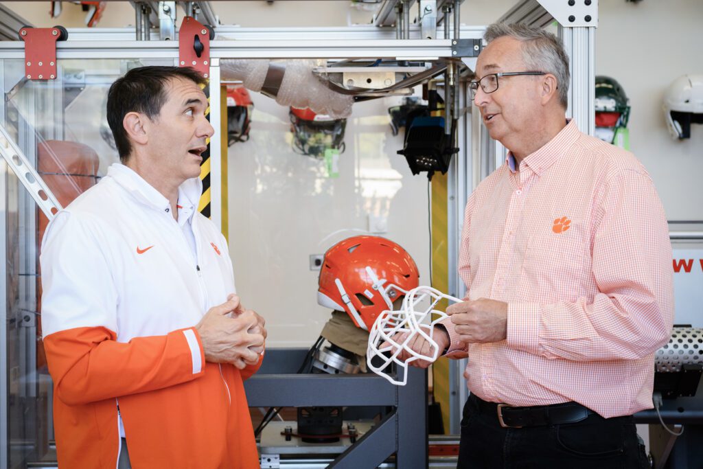 Two men stand on either side of a glass box that holds a football helmet
