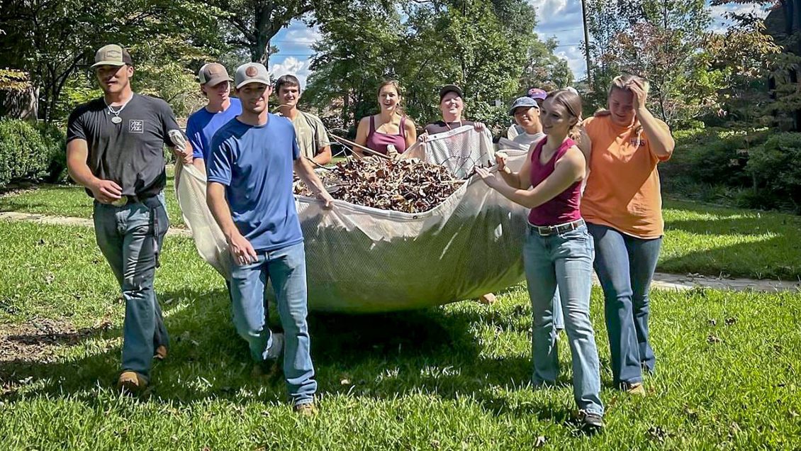 A group of nine young people carry a tarp full of leaves and sticks through a grassy lawn.