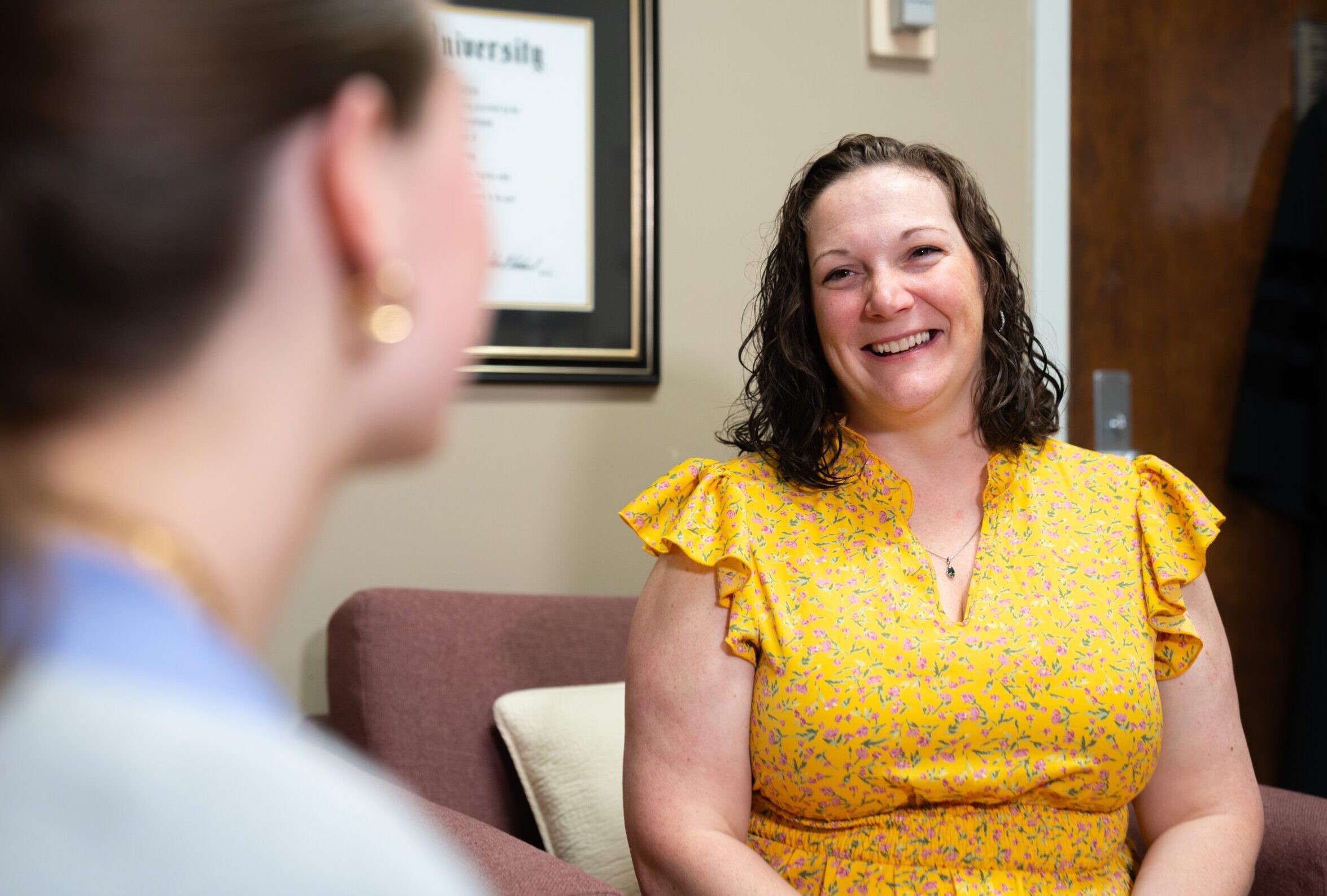 a smiling woman in a yellow dress sits in a chair facing a student in an office setting