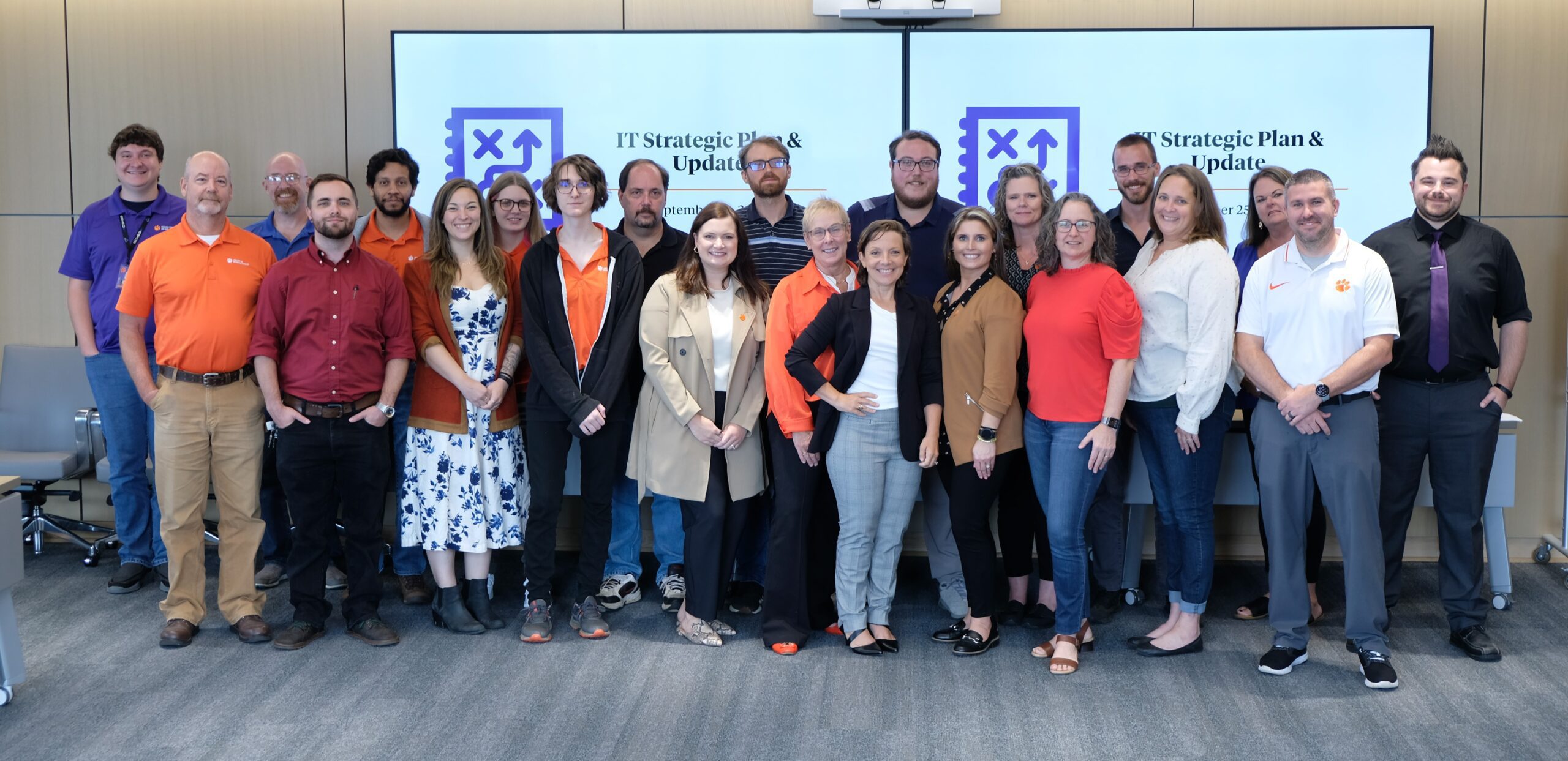 A group of CCIT employees stands in the middle of the Executive Boardroom in the Powers College of Business in front of two screens with white backgrounds.