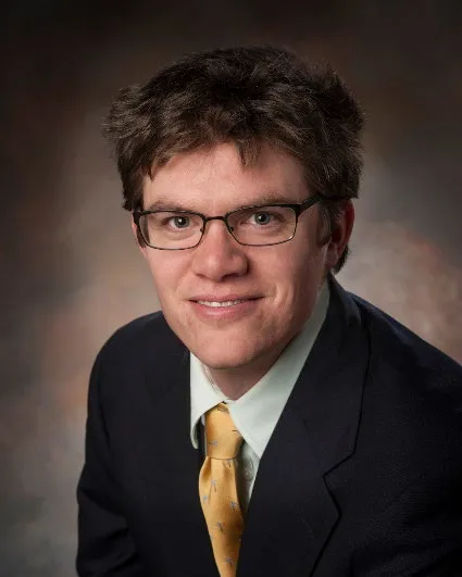 A man wearning glasses and a black suit with a yellow tie.