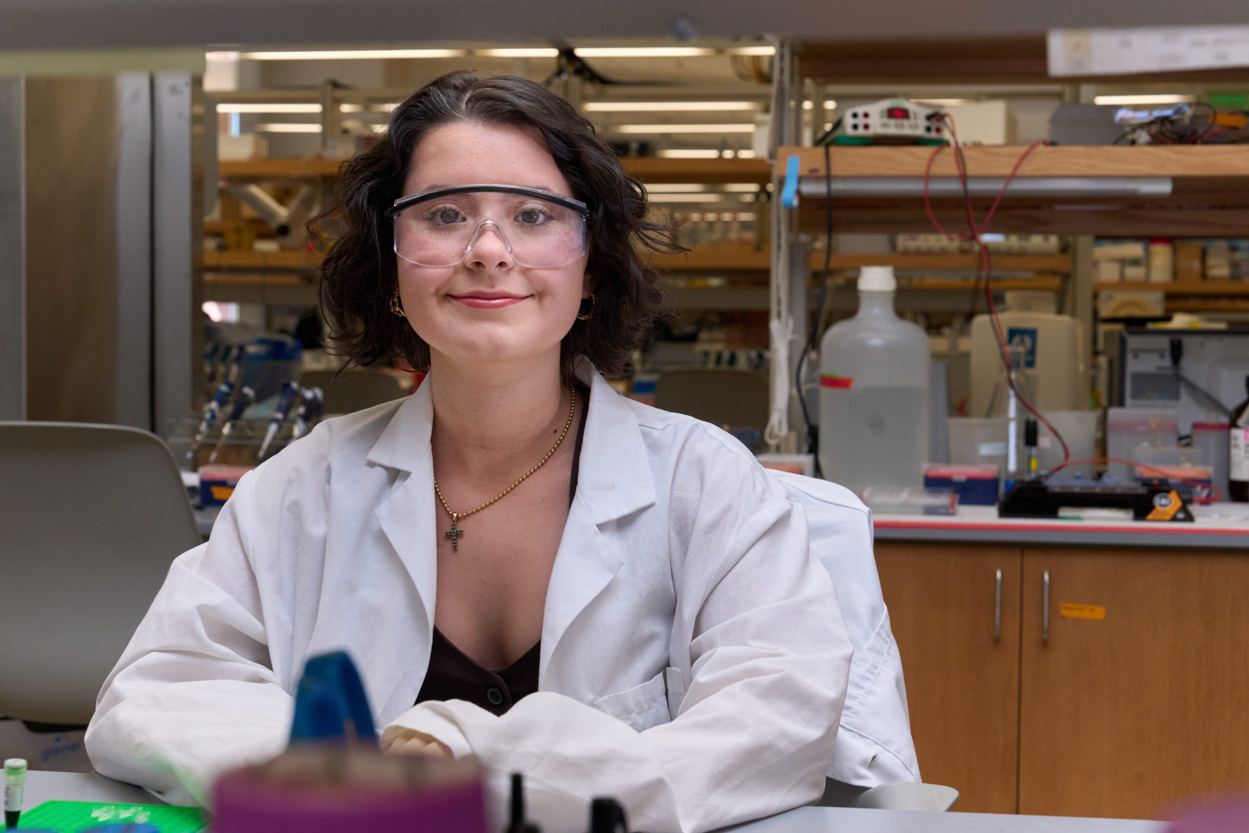 A woman wearing a white lab coat sits at the bench in a science lab