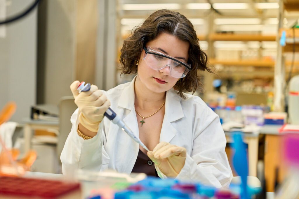 Woman wearing goggles and a white lab coat pipettes in a. lab