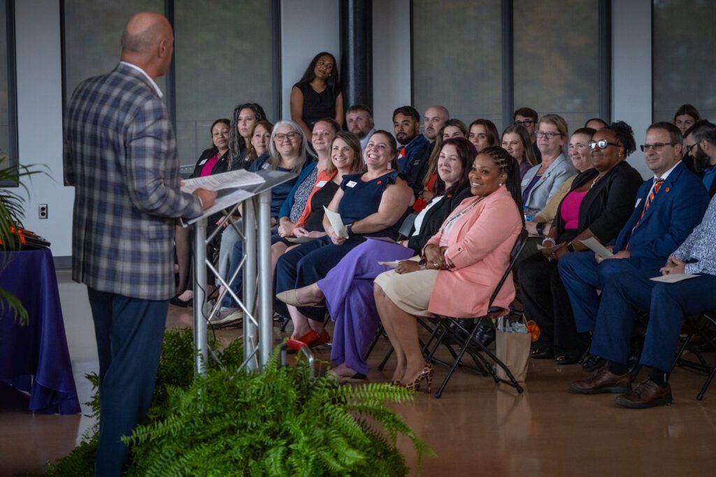 A man in a plaid suit jacket stands at a podium and speaks to a group of individuals seated in chairs.