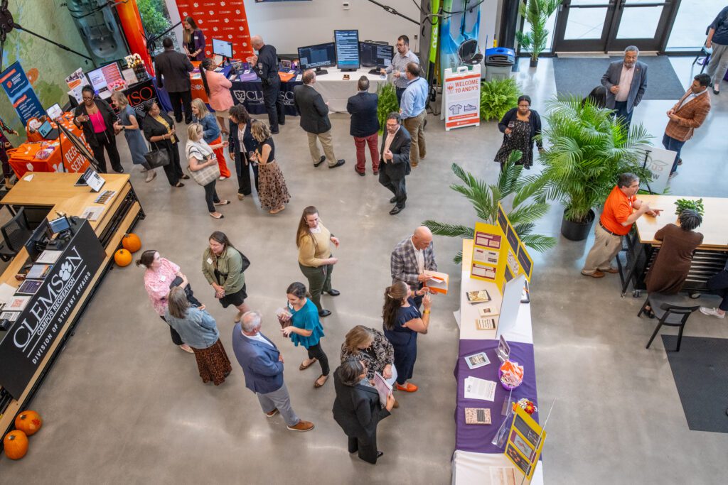 Individuals mull around an atrium with cement floors where long tables are set up with posters and presentations.