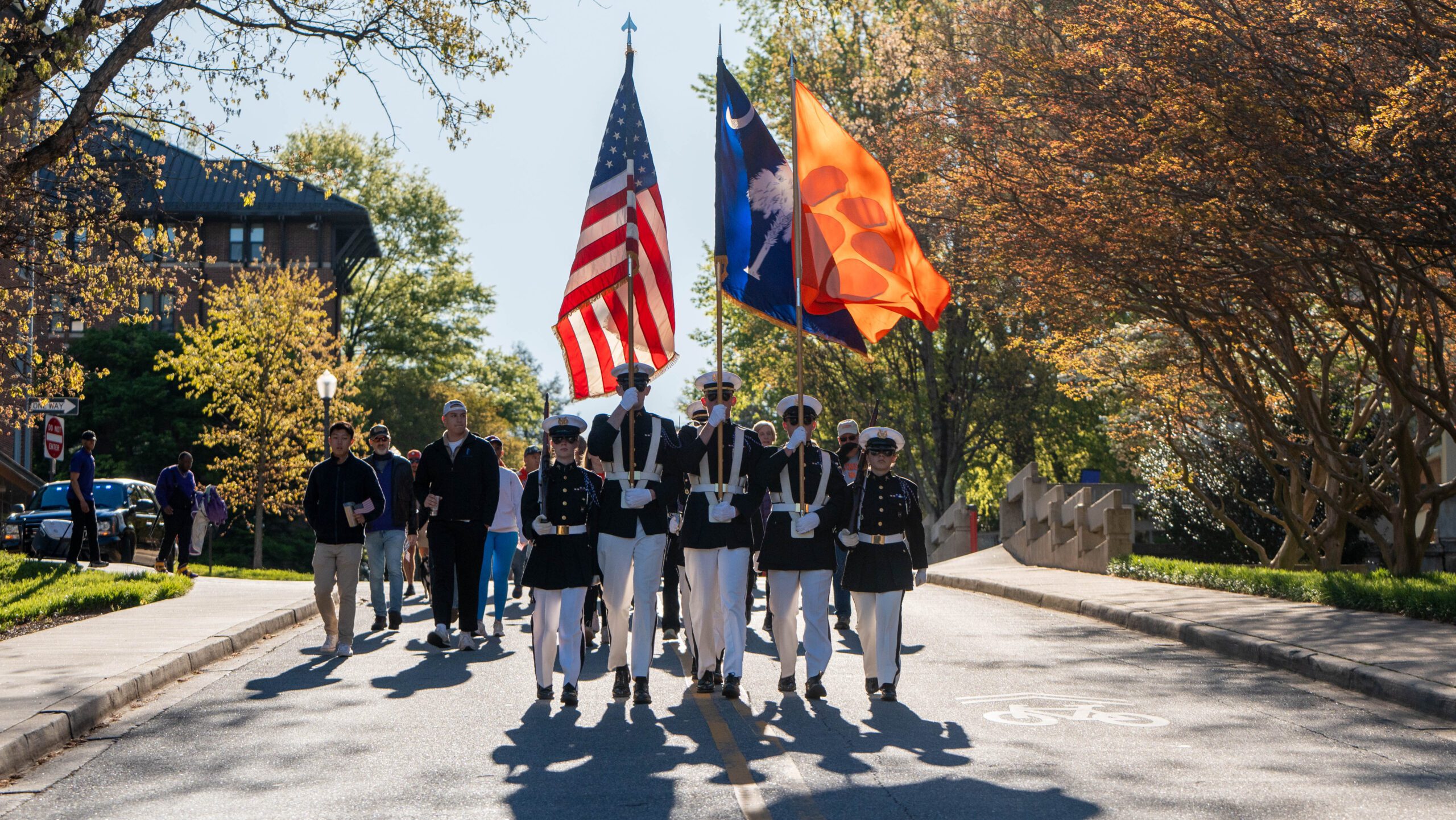 Three students in military uniforms walk through the street holding an American flag, South Carolina flag and Clemson flag.