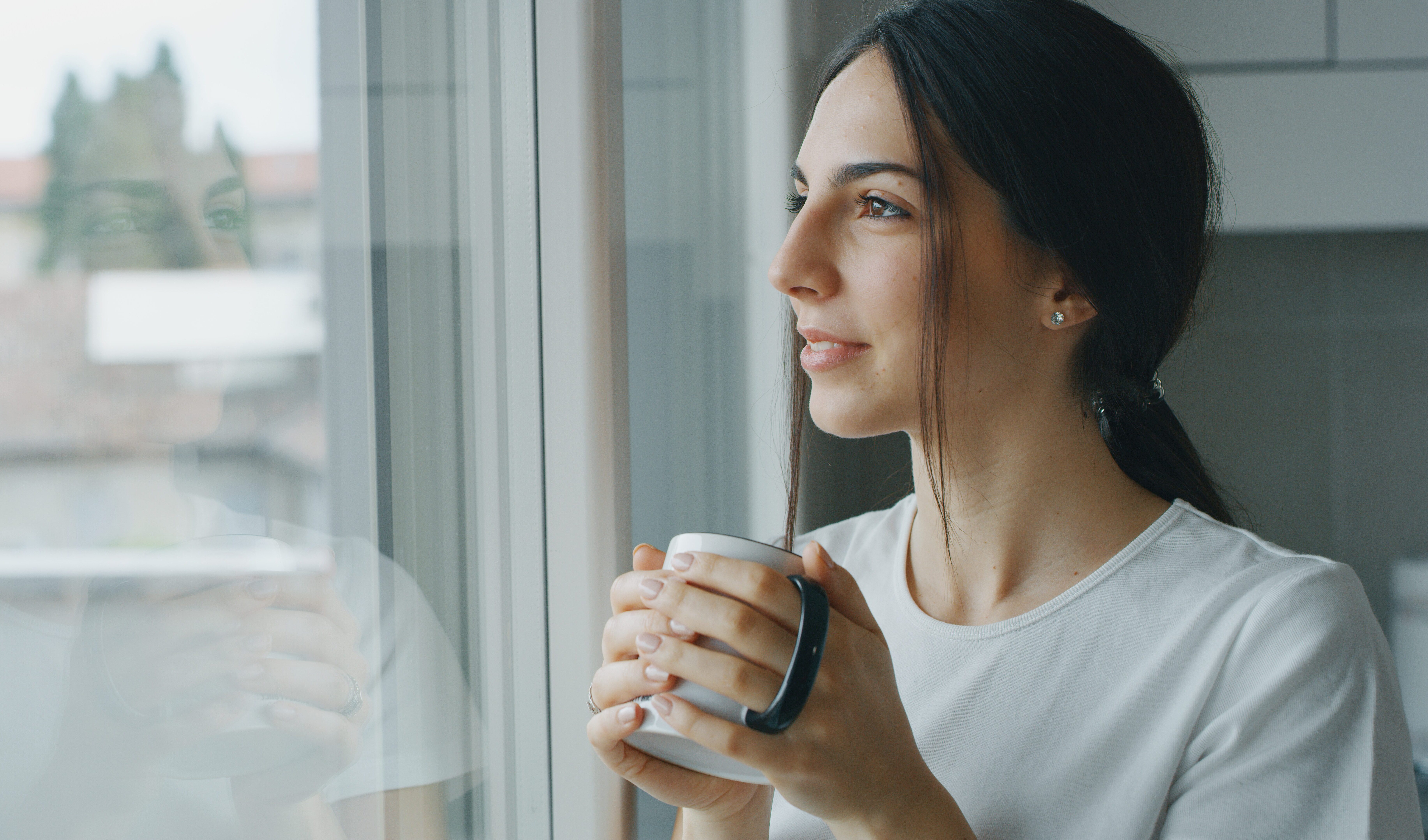 Portrait of a pensive young woman is drinking a tea and looking through the window in the morning in the kitchen