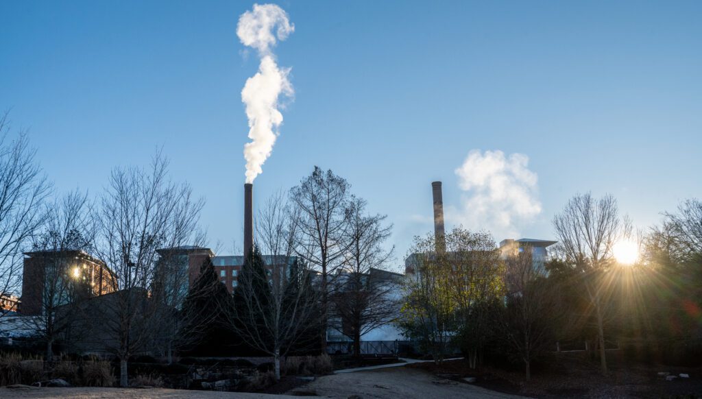 Steam spews into the sky from a tall brick smokestack as the sun flares through the branches of the trees in front of it.