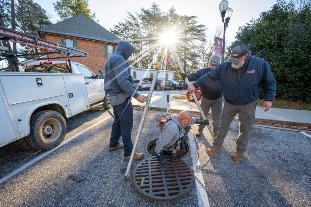 Two men hold a tripod as another man climbs out of a manhole at their feet, with the sun shining straight into the camera making it look like a flare is on top of the tripod (like a Christmas tree)