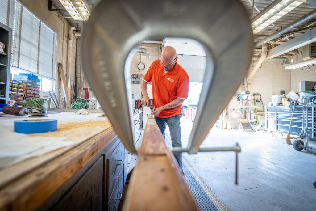 A man is seen through a metal clamp running a sander over a board