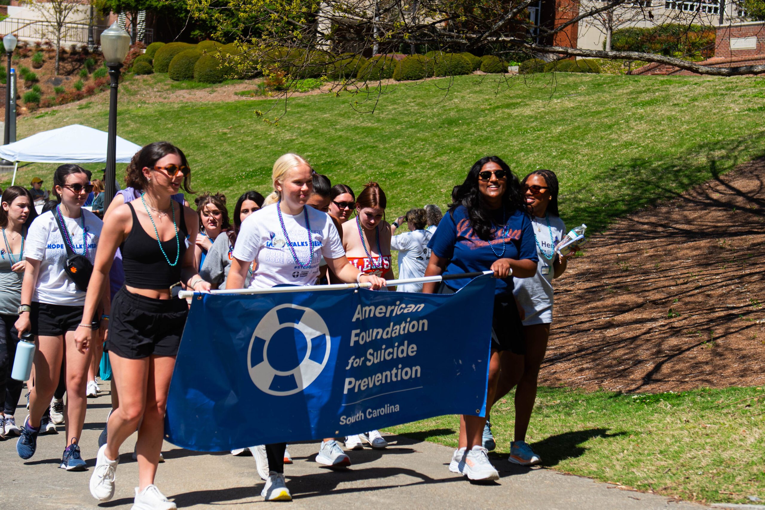 three young women are walking and carrying a blue American Foundation for Suicide Prevention South Carolina banner as a group of people walk behind them
