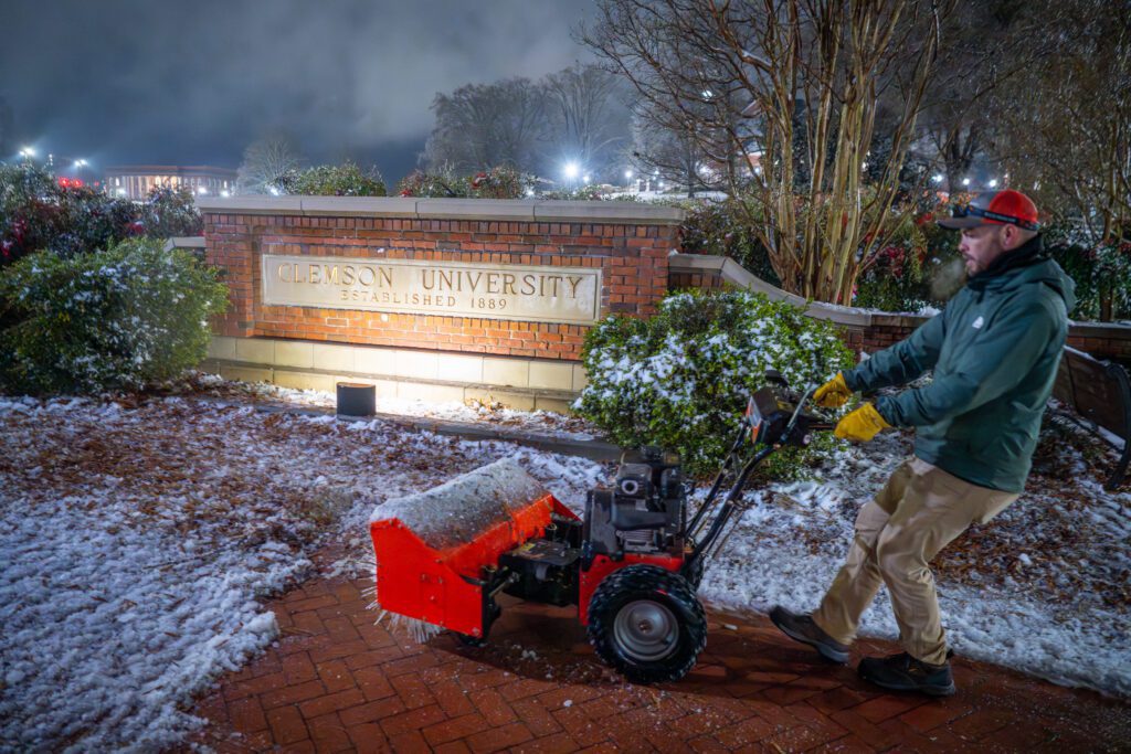A man uses a snow blower to clear the sidewalk in front of the Clemson University sign at night