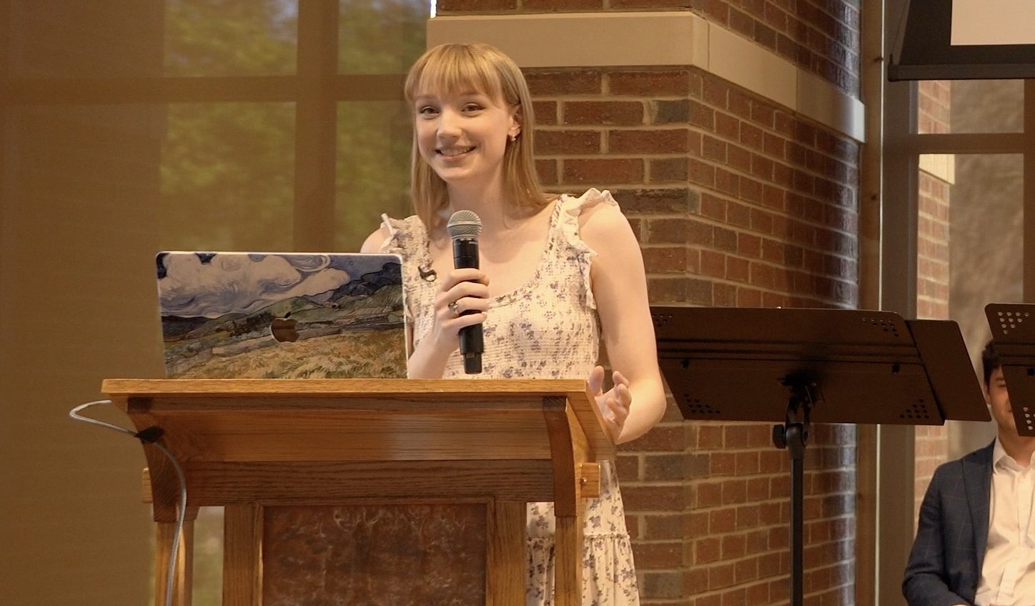 A young woman holding a microphone stands and smiles behind a podium.